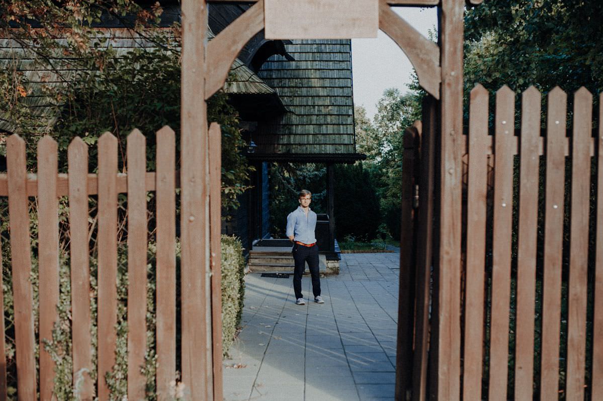 blonde man in blue shirt brown belt and navy trainers stands beside wooden entrance to Stefan żeromski museum in nałęczów